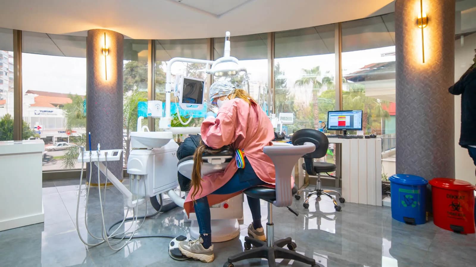 Een behandelaar met mondmasker bij Ballıpınar Dental behandelt een patiënt in een lichte behandelkamer, beiden van achteren gezien